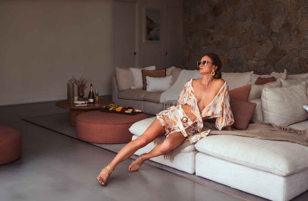 A person in a floral outfit and sunglasses, looking effortlessly chic at forty, lounges on a sofa in a modern, stylish living room with stone walls and a round coffee table.